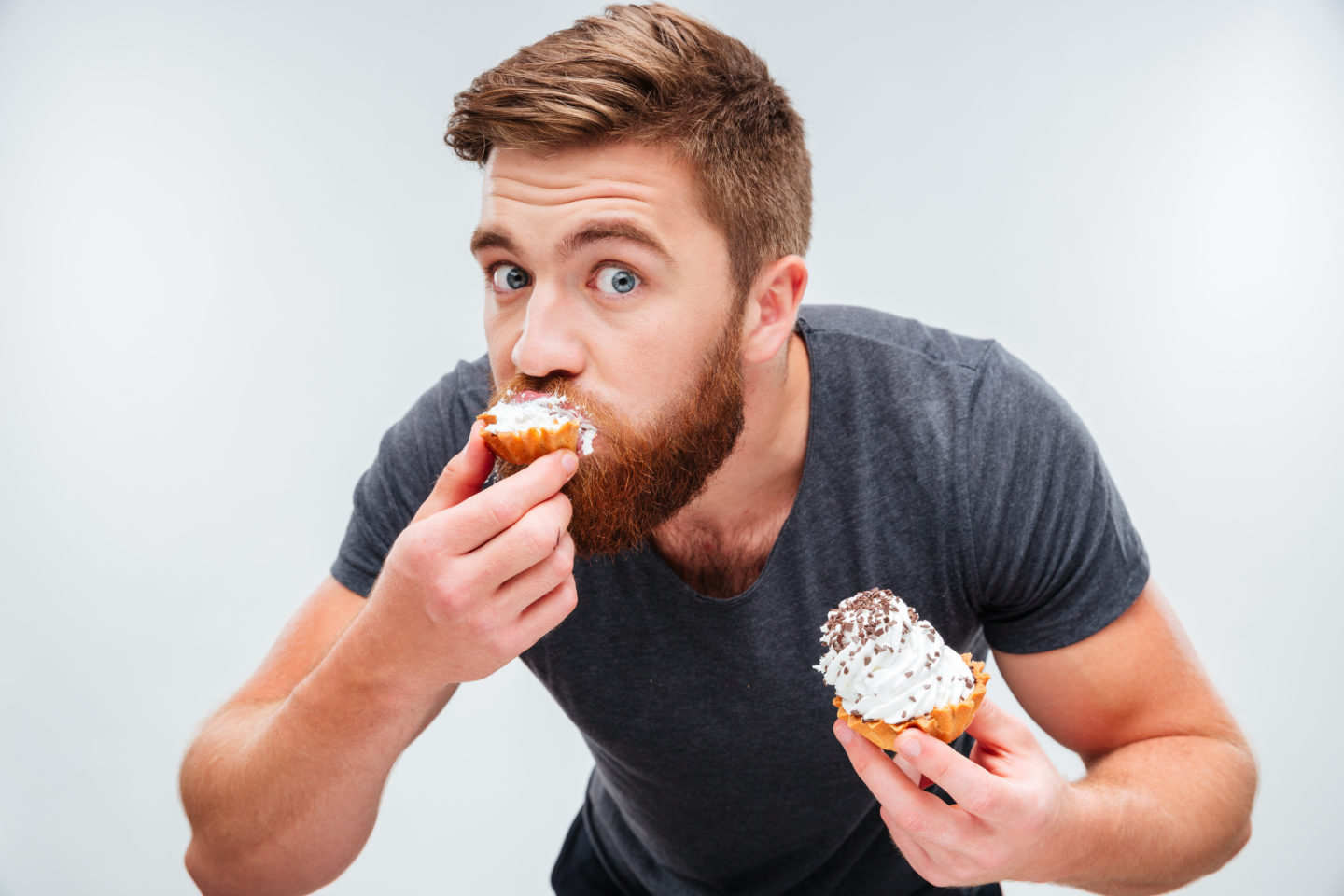 Close up portrait of a hungry bearded man biting cream cake isolated on white background